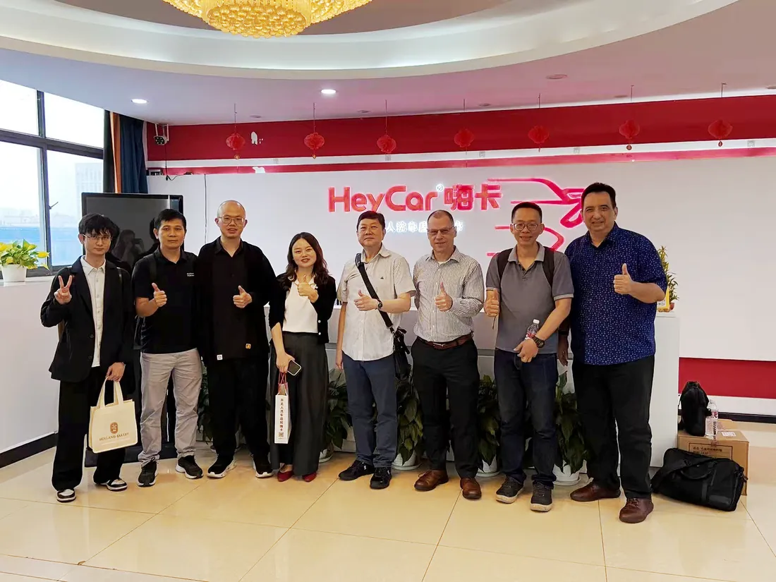 A group of eight people standing together in front of the illuminated red HeyCar sign in the company's reception area, most of them giving a thumbs-up.