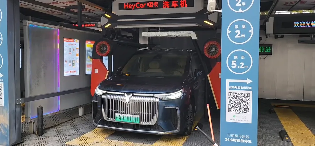 A dark SUV inside a HeyCar automatic car wash bay, with the washing gantry overhead, red drying fans on either side and a blue pillar listing the two metre width and 5.2 metre length limits.