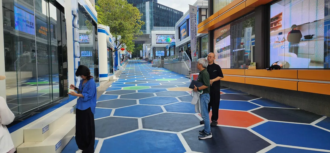 An open-air retail street at Robotmall in Shenzhen, with a hexagon-patterned floor in blues and orange, large video screens in the shopfronts on both sides and visitors standing in conversation.