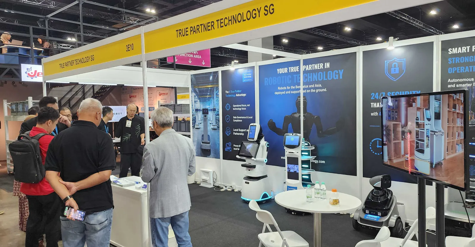The True Partner Technology stand at the trade fair, with a reception robot, a delivery robot and a security robot in front of the stand's display panels.