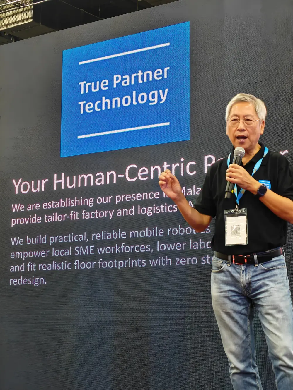 Vincent Lum speaking with a microphone in front of a large presentation screen carrying the True Partner Technology logo.